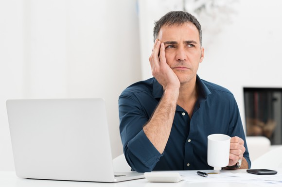 A worried man holding his head in his hand and pondering the future in front of his laptop.