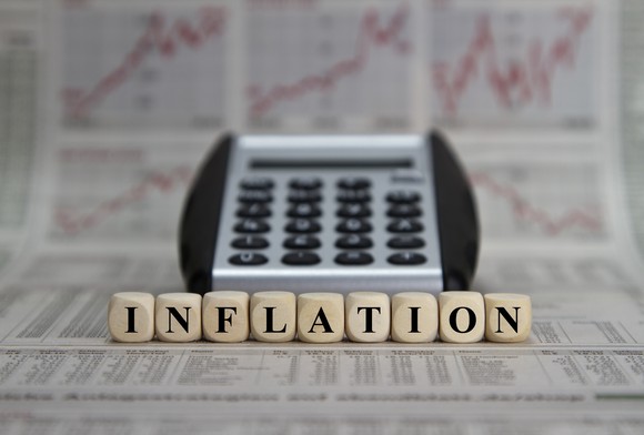 The word "inflation" spelled out with dice in front of a calculator and various rising charts in a newspaper.