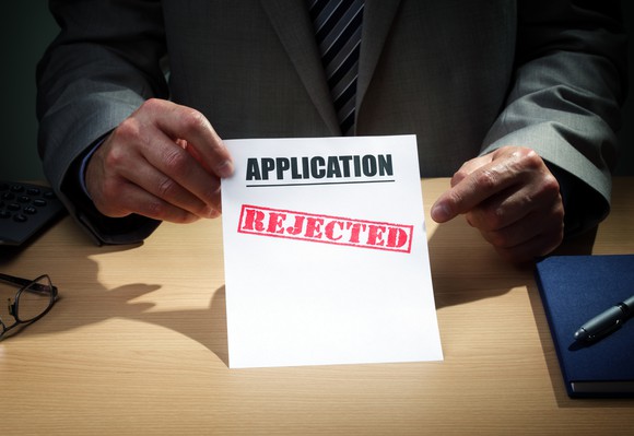 Man in suit holding up a piece of paper that says "Application" with the word "rejected" stamped on it.