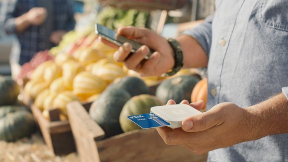 Man holding Square card reader in one hand and a smartphone in other hand with bins of produce in background.