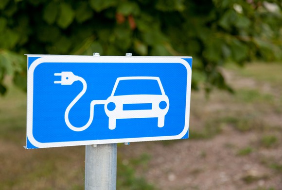 Outdoor sign with an image of a car with an electrical cord attached to it -- concept for an electric car. Sign indicates a charging station.