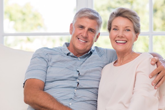 A smiling older man sits on a sofa next to a smiling older woman, with an arm around her shoulder.