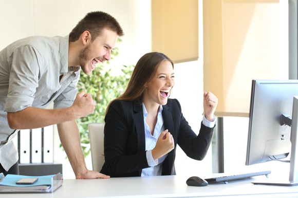 Smiling and making fists, a man and woman celebrate while looking at a computer screen.