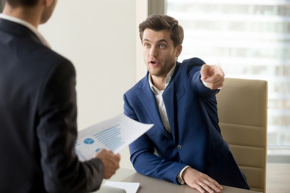 Man in suit pointing toward the door while meeting with another man holding a document
