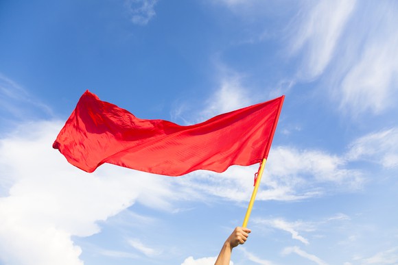 An arm holding up a red flag against a blue sky.
