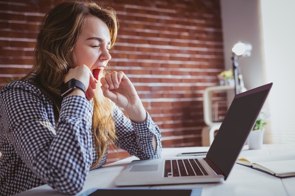 A young woman yawns in front of a laptop.