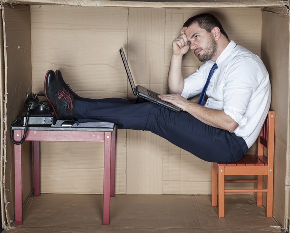 Young businessman with a laptop and a desk phone, sitting on a small chair inside a large cardboard box.