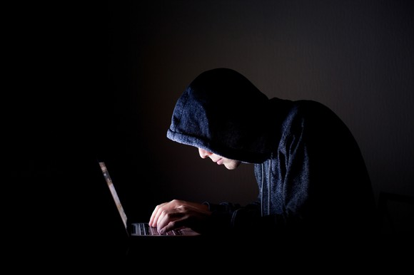 A person in a black hoodie in a dark room, hunched over a laptop with the only light coming from the computer's screen.