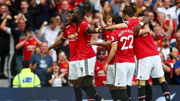The Manchester United team celebrates on the field after defeating a rival. MU's team wears red jerseys and white shorts.