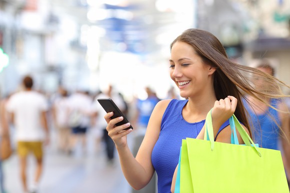 Smiling woman with shopping bags standing in street, looking at mobile device.