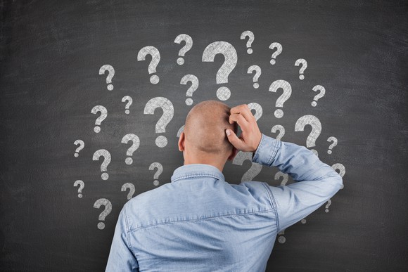 Man with hand to head facing chalkboard with lots of question marks drawn on it