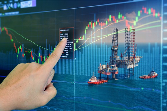 A hand pointing at a stock screen with an oil drilling rig in the background.