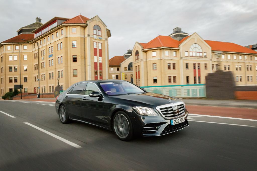 The Mercedes-Benz S Class Salon in the color black is shown speeding on a road with buildings in the background.