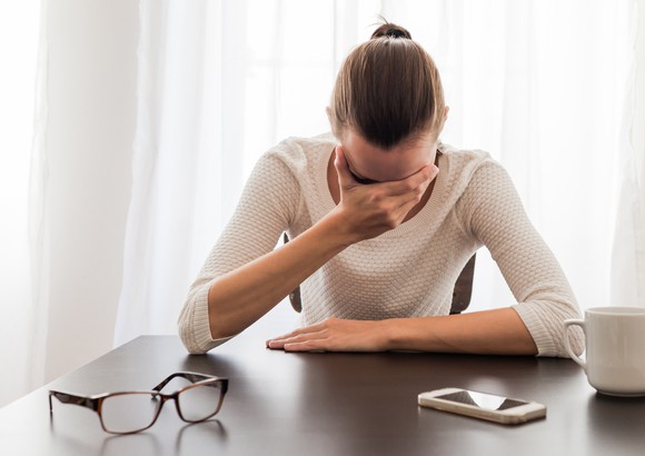A woman sits at a table, glasses and phone lying in front of her, as she holds her face in her hand, appearing to be crying.