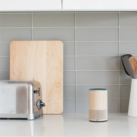 An wooden-looking Amazon Echo on a kitchen counter with toaster, cutting board and mixing spoons.