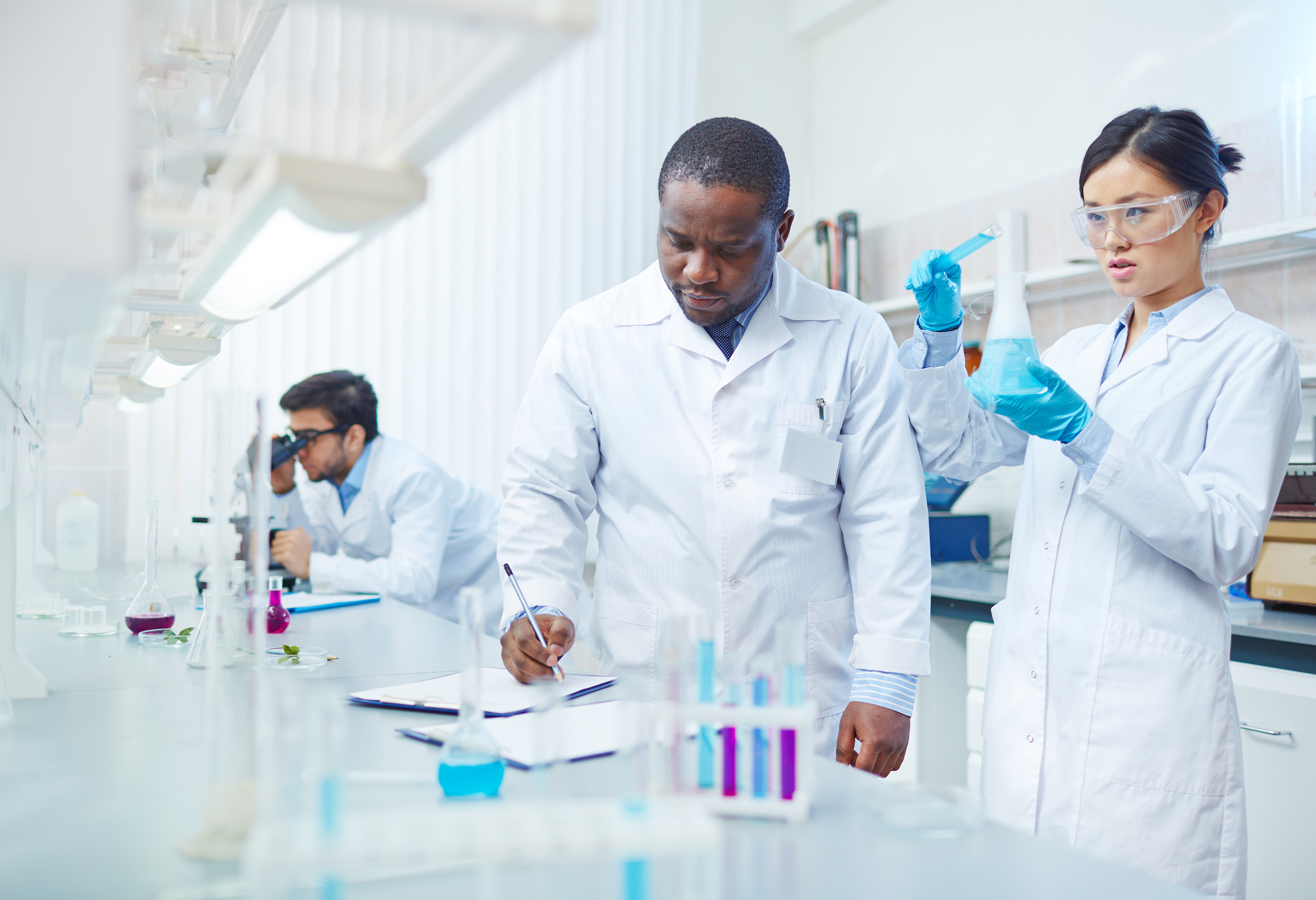 Three scientists in a lab, with one writing on a notebook, one holding a beaker, and another in the back looking through a microscope.