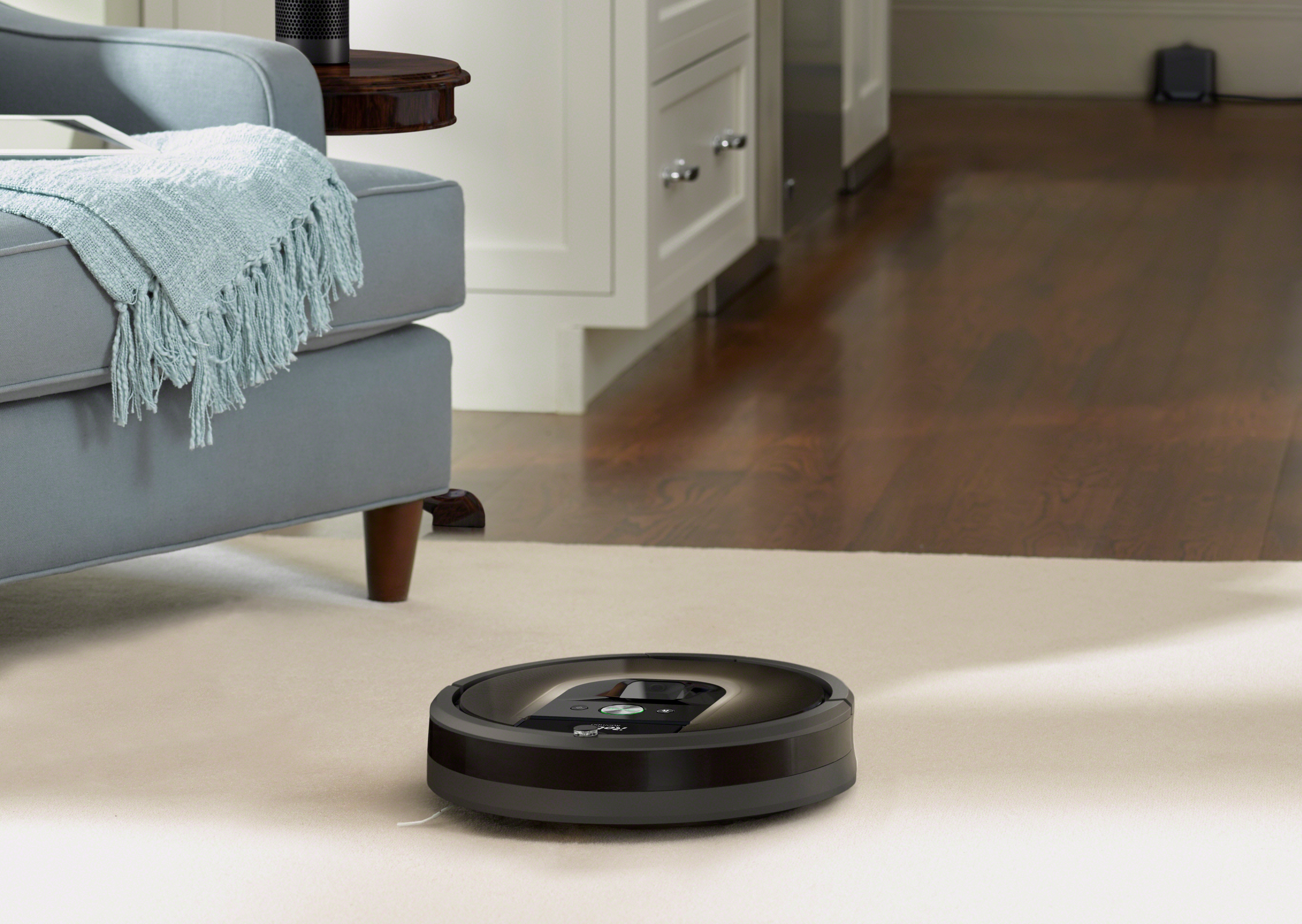 A Roomba vacuuming an area rug on a hardwood floor in a room with a blue armchair.
