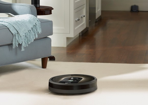 A Roomba vacuuming an area rug on a hardwood floor in a room with a blue armchair.