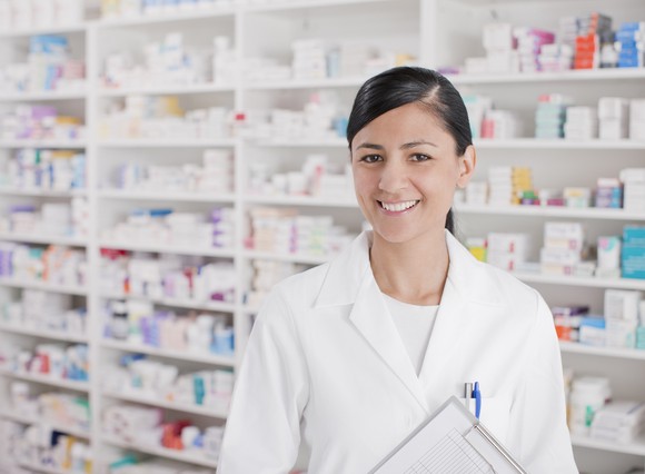 Pharmacist in a white lab coat in front of a set of white shelves with pharmaceuticals on them.