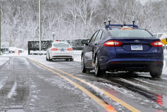 Two Ford Fusion sedans with visible self-driving hardware are shown driving on a snowy road.