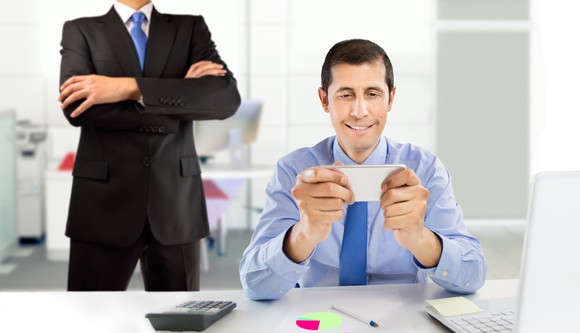 A professionally dressed male checks his phone while a man in a suit and tie stands behind him with arms crossed