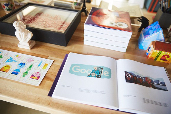 Desk at Google with book open to various Google doodles