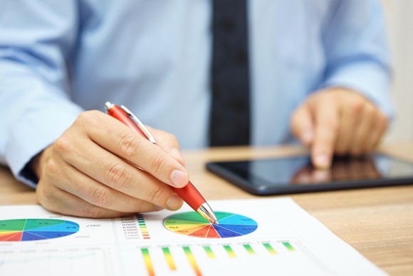 A man standing at a desk touches a tablet with one hand and points a pen to a pie chart with the other.