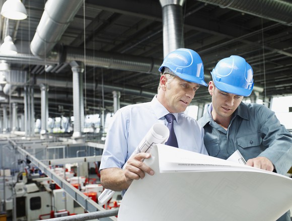 Two men in blue hard hats review documents on a factory floor