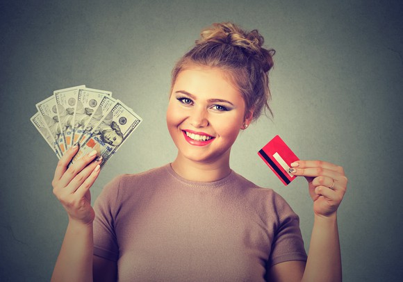 smiling young woman holding credit card in one hand and hundred dollar bills fanned out in the other