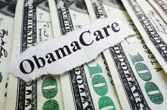 A piece of paper that reads Obamacare lying atop a fanned pile of hundred dollar bills.