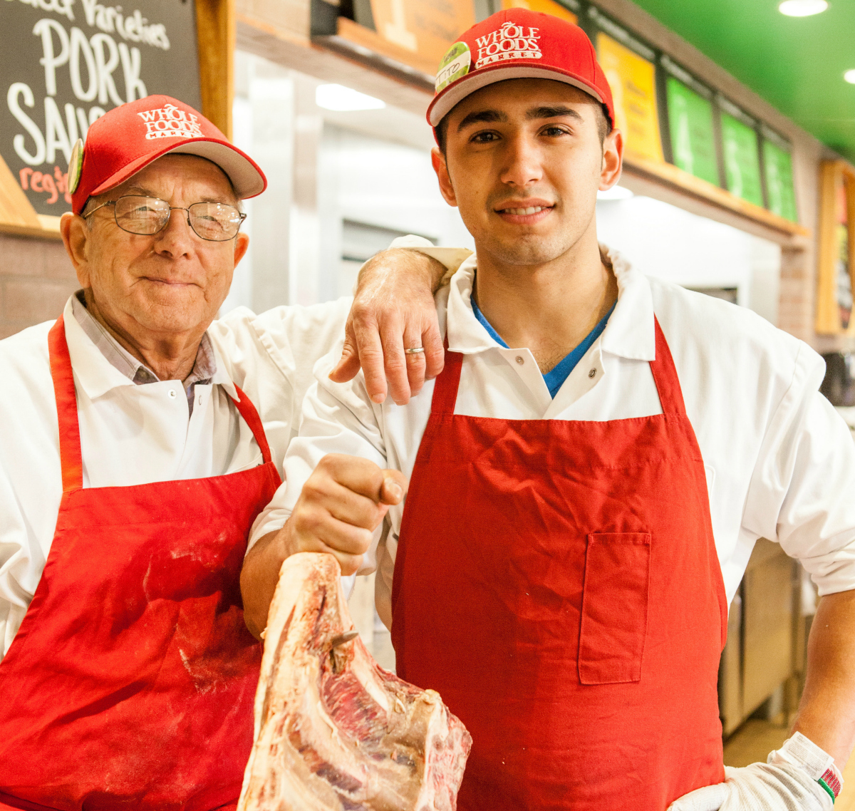 Two workers wearing red Whole Foods hats and red aprons stand together at a Whole Foods.