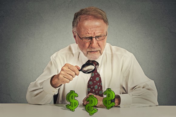A senior man using a magnifying glass to examine dollar signs on a table.