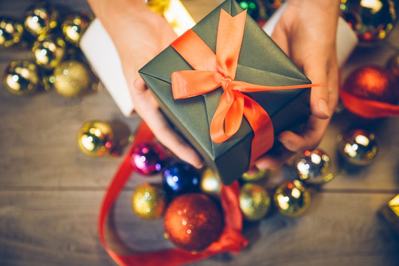 A person holds out a small gift box with Christmas ornaments in the background.
