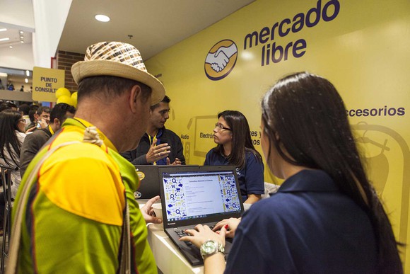 MercadoLibre employee working with customer in front of a computer display screen, with logo in background.