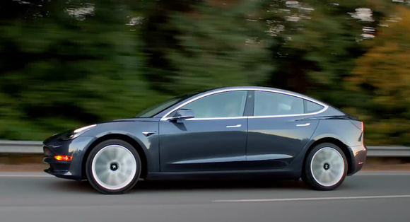 Side view of a black Model 3 driving on road with fall greenery in background.