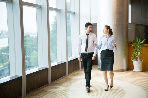 Businessman and businesswoman walking by large office windows discussing a matter.