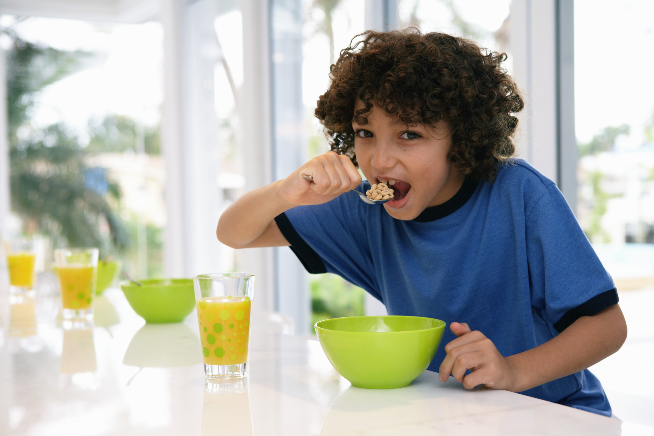 A boy in a blue shirt leans over a green bowl on a counter as he lifts a spoonful of cereal to his mouth.