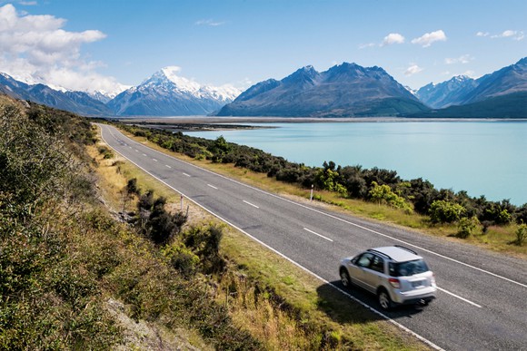 Car driving along a glacial lake in New Zealand, with relatively clear skies against a mountain backdrop.