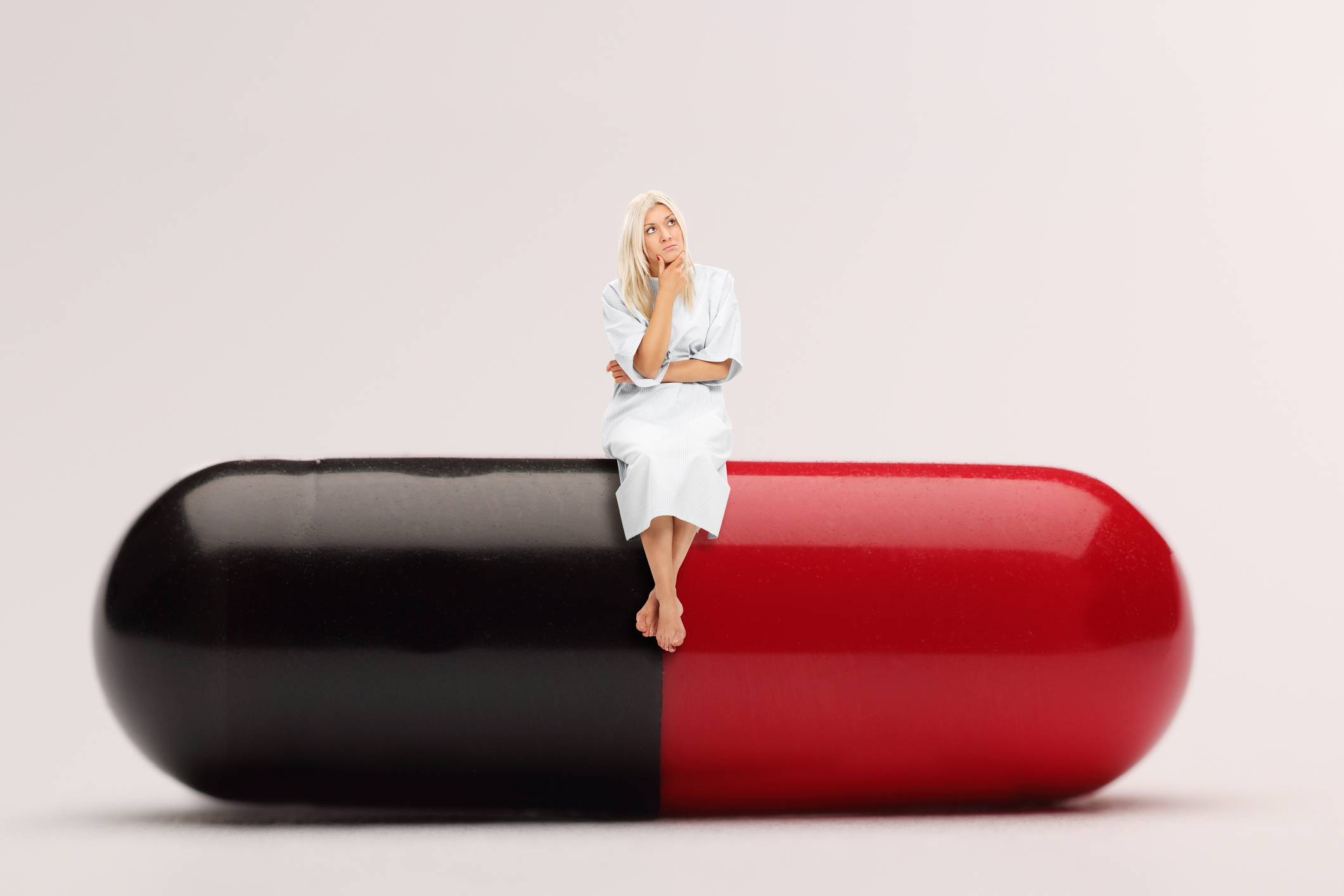 Person in hospital gown sitting on top of a giant red and black medicine capsule.