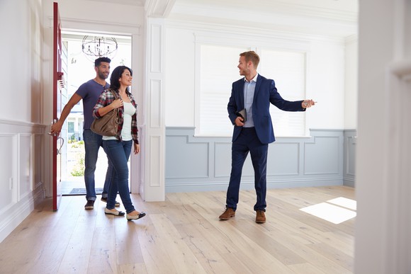A man in business attire gestures to a couple walking into an empty house.