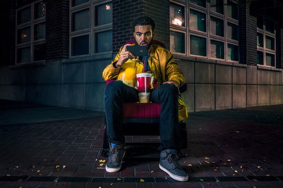 A man sitting on a chair with a tub of popcorn and a large drink looking at a Razer Phone.