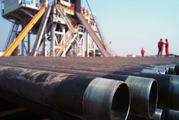 A close-up of drill pipes with oil workers and a rig in the background.