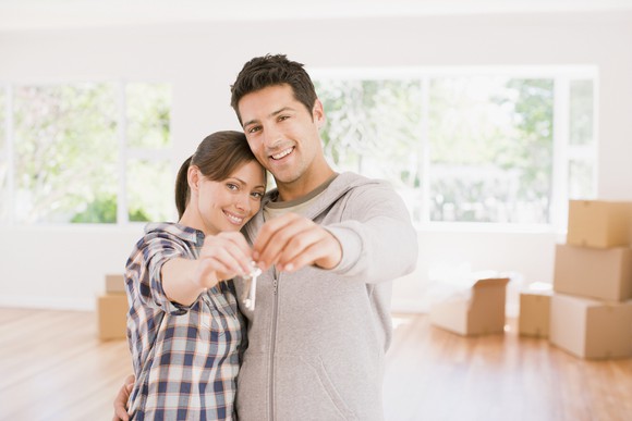 A couple holding a key to their new home, with moving boxes in the background.