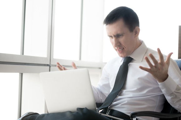 A frustrated man holding his hands up and reading something on his laptop.