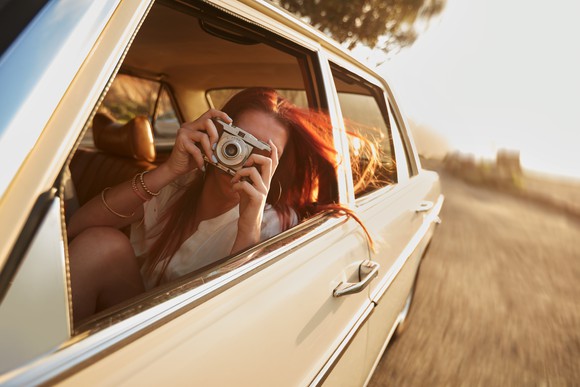 Woman taking a photo on vintage camera (pointed at viewer) on a rural road trip.