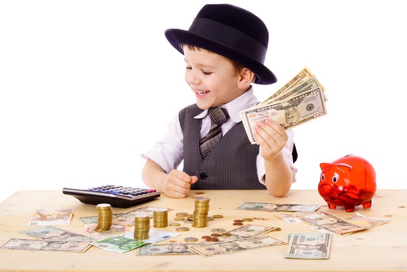 A kid in a tie and hat holds a wad of cash and looks at money on the table.