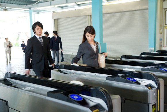 Two young Japanese women in business attire cross through a train-station turnstile.