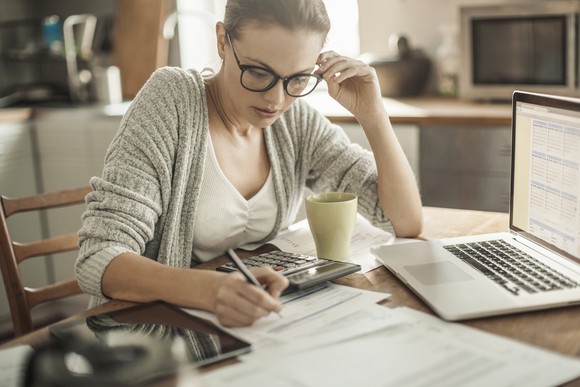 A woman at a kitchen table uses a calculator and a computer.