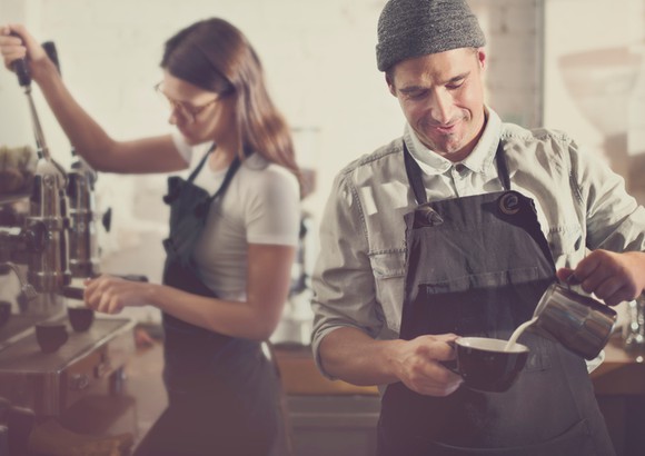 Two aproned baristas working with focus in small coffee shop.