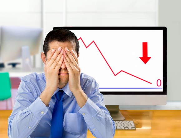 Man covering his face in front of a computer screen with a downward graph and an arrow pointed downward